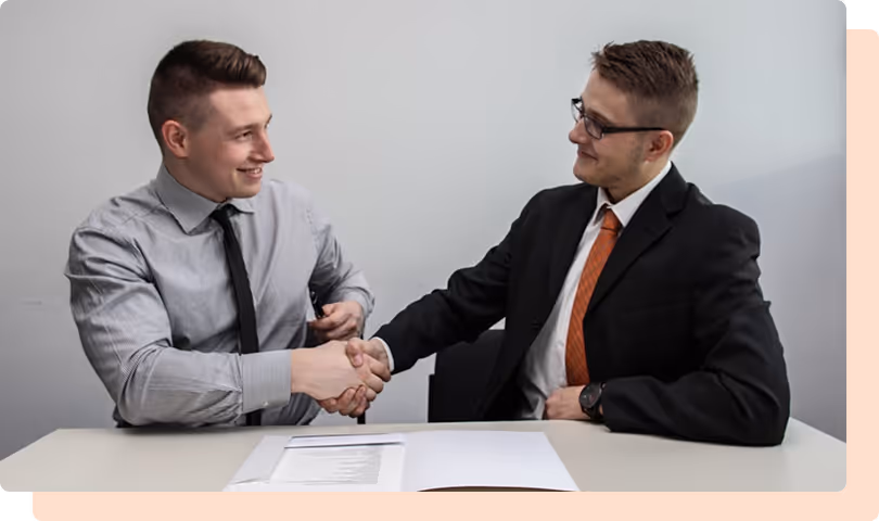 Two men in business attire shaking hands over a table with documents, smiling at each other.