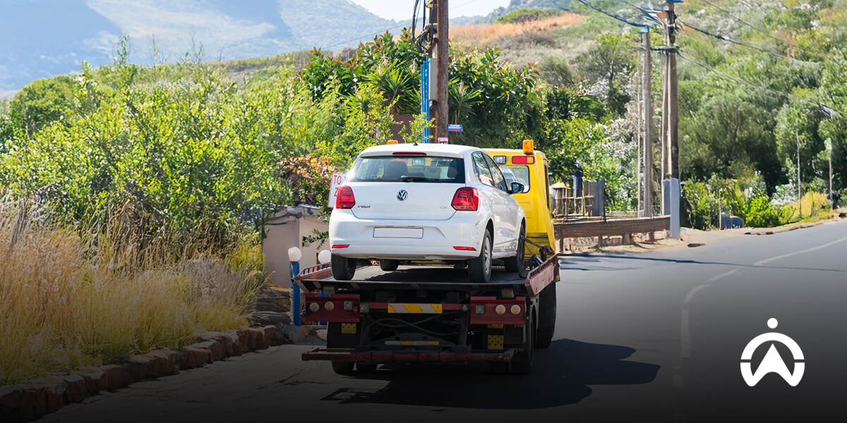 A white car on a tow truck on a roadside with greenery in the background.
