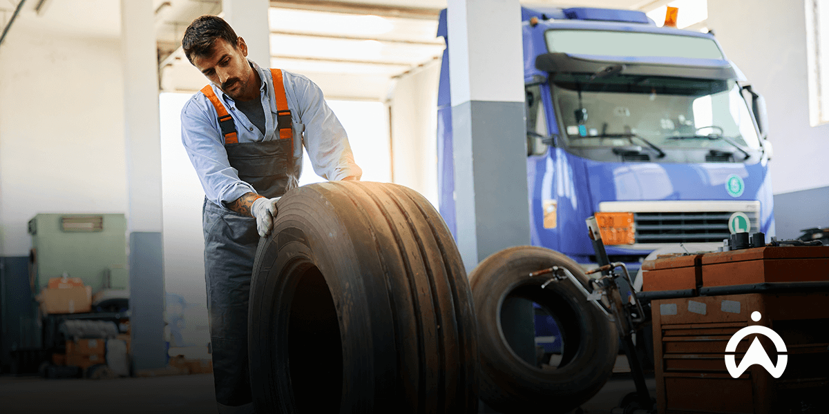 Mechanic in overalls rolling a large tire with a blue truck in the background.