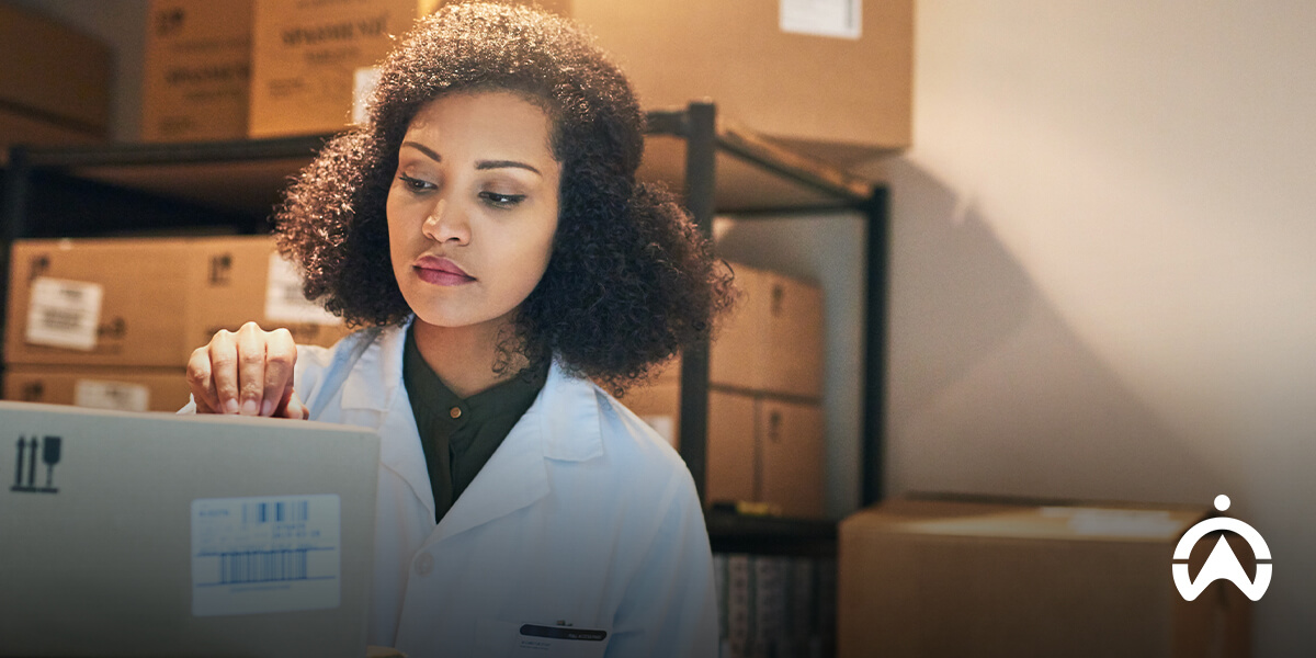 A pharmacist in a white lab coat handles a cardboard box in a dimly lit storage area filled with stacks of boxes.