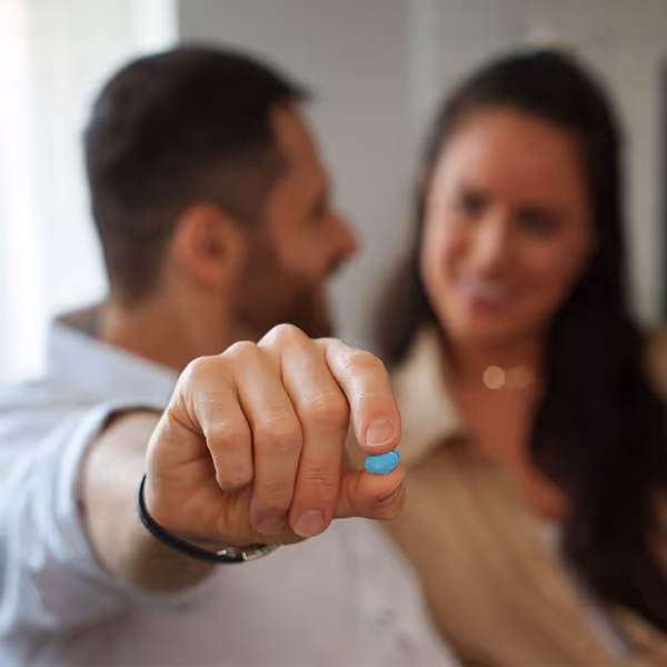 Man holding a blue pill between thumb and forefinger with a woman smiling in the background.