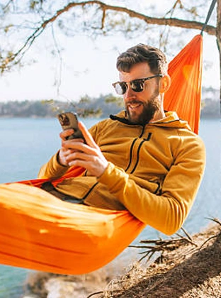 Man wearing sunglasses and a mustard jacket relaxing in an orange hammock near water while using a smartphone.
