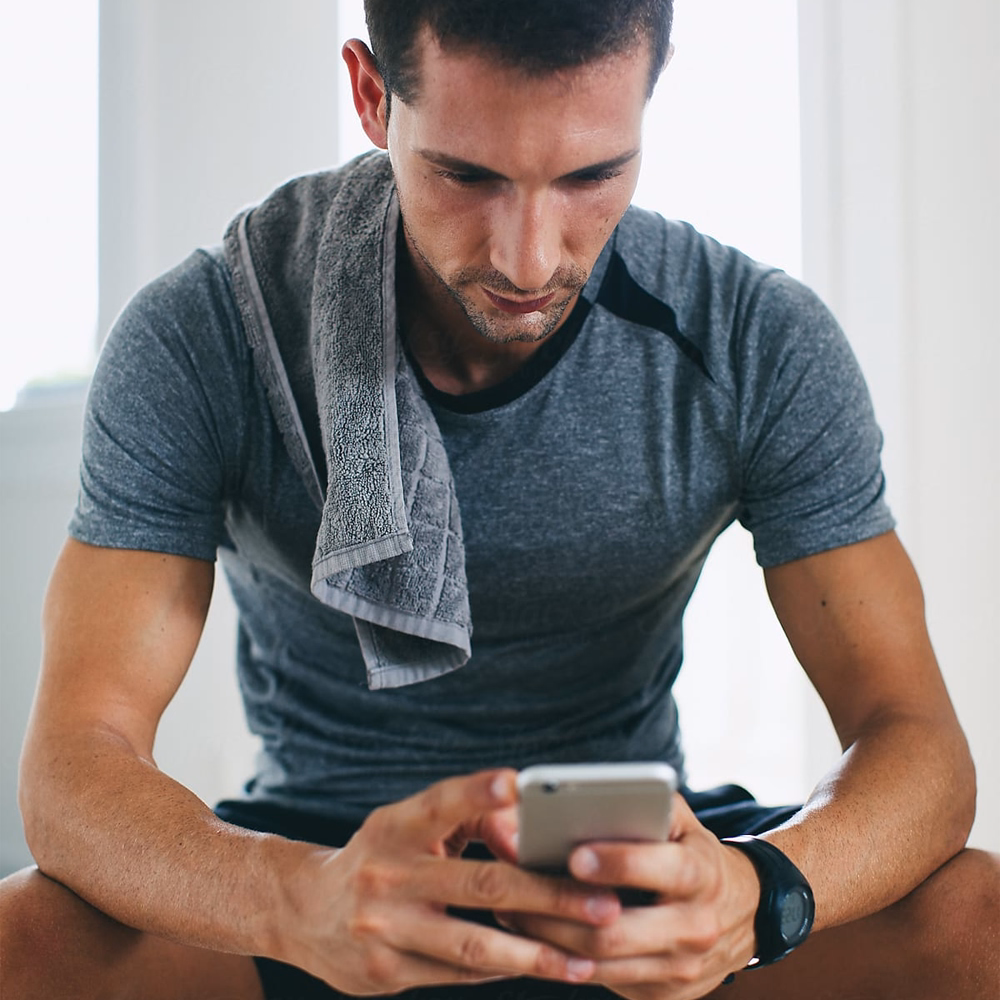 Athletic man with a gray towel over his shoulder looking at his phone, sitting indoors.