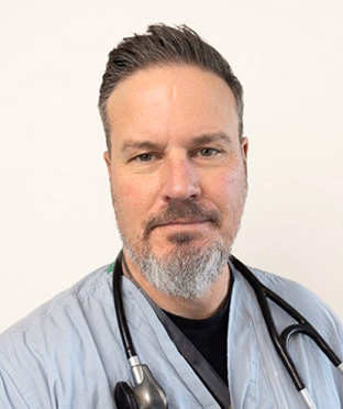 Portrait of Roger Firsoff, a man with short dark hair and a gray beard wearing light blue medical scrubs and a stethoscope, looking at the camera.