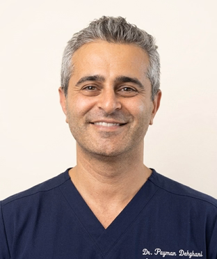 Portrait of Dr. Payman Dehghani, a smiling man with short gray hair wearing navy medical scrubs, facing forward against a plain background.