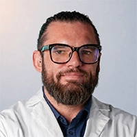 Portrait of Dr. Mark Broussenko, a man with dark hair pulled back, a full beard, and glasses, wearing a white lab coat over a blue shirt.
