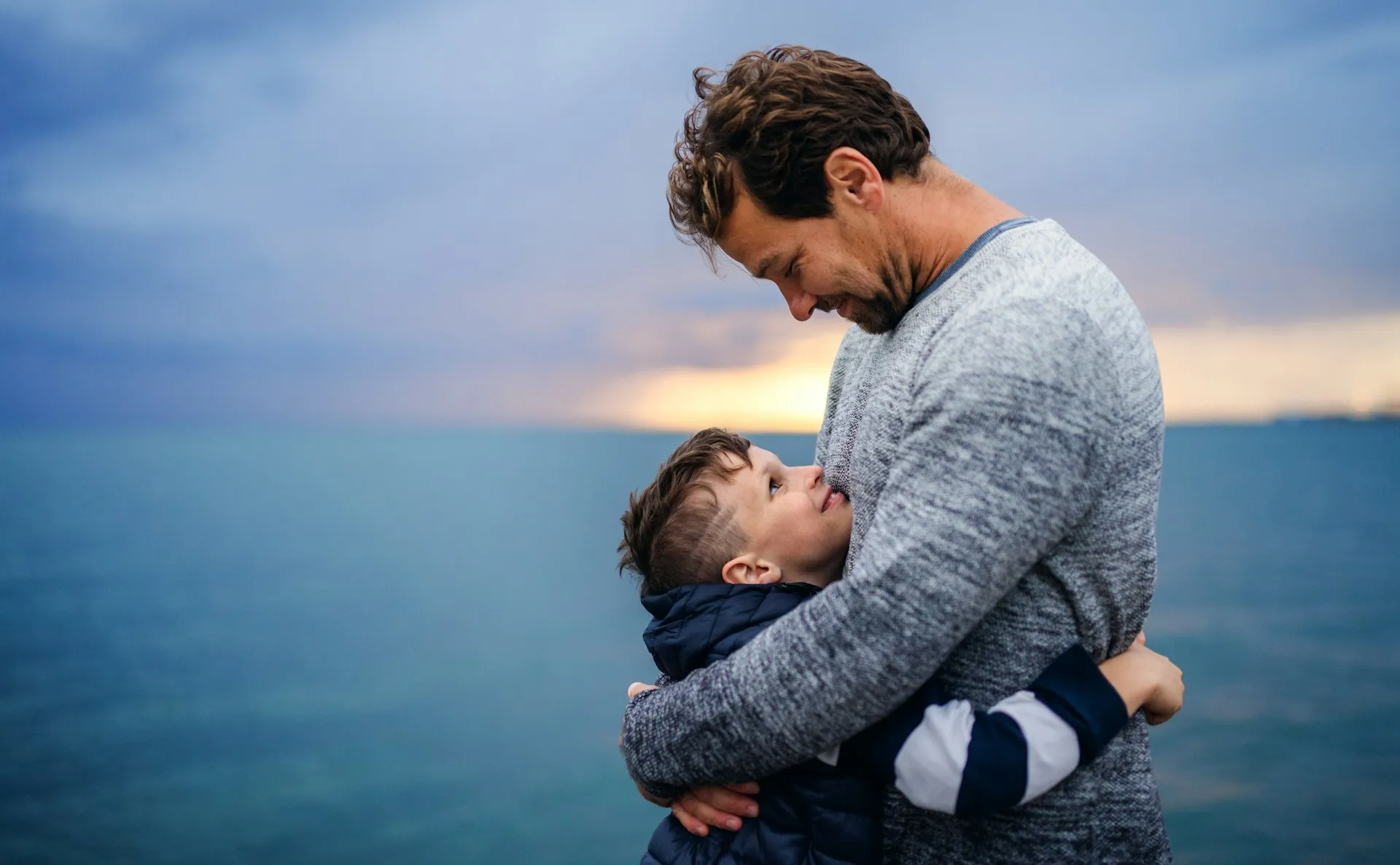 Man and young boy embracing by a body of water at sunset, sharing a tender moment with the boy looking up at the man.
