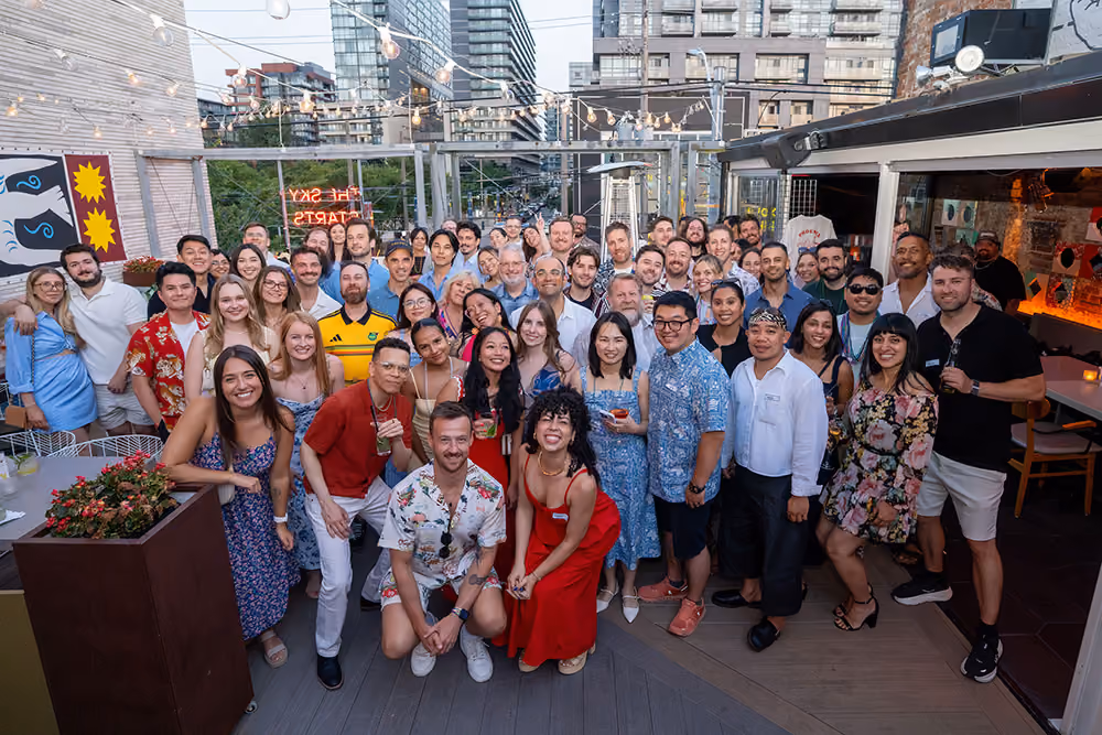 Large group of diverse people smiling and posing together on a rooftop patio with string lights and city buildings in the background.