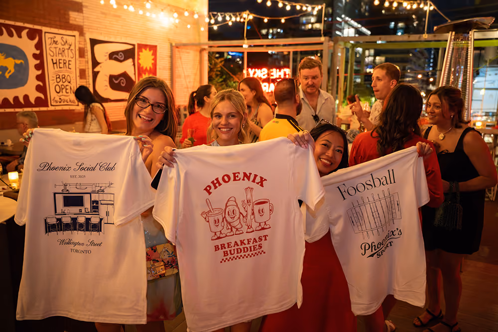 Three young women at an outdoor evening gathering hold up white t-shirts with different prints: the left shirt shows 'Phoenix Social Club' with a kitchen design, the center shirt says 'Phoenix Breakfast Buddies' with cartoon breakfast items, and the right shirt features a foosball table graphic labeled 'Foosball Phoenix's'. People and string lights decorate the lively background.