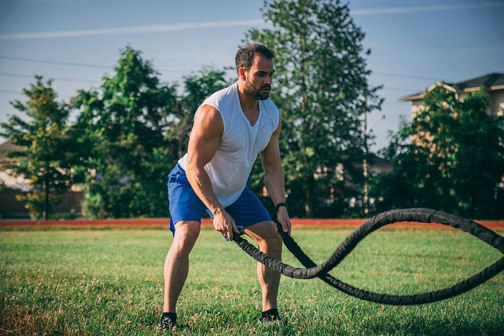 Man exercising outdoors on a grass field, using battle ropes in a bent stance on a sunny day.