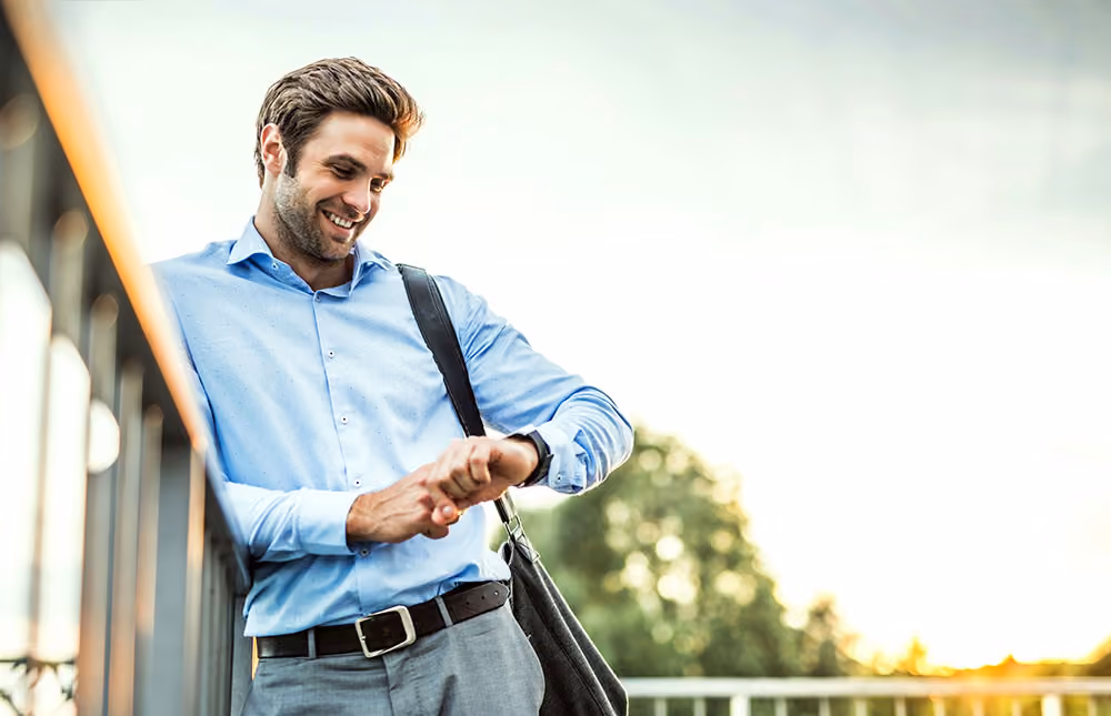 Man in a blue dress shirt checking his watch while walking outdoors at sunset