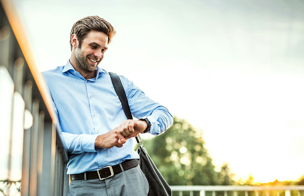 Man in a blue dress shirt checking his watch while walking outdoors at sunset