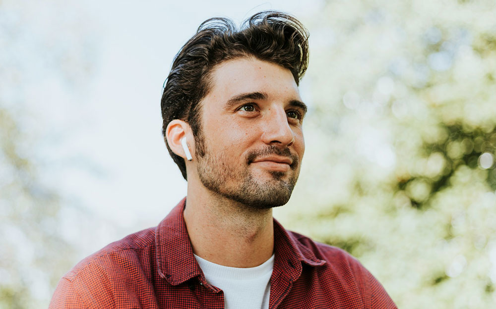 Man in a red checked shirt with a wireless earbud looking off camera outdoors