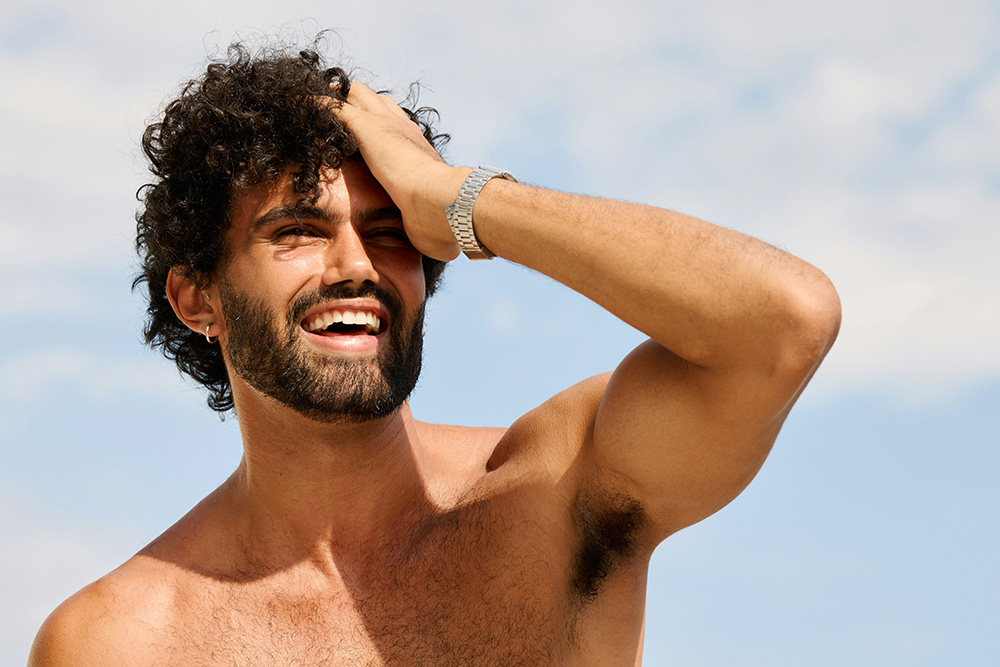 Smiling man with curly dark hair and a beard shading his eyes with his hand on a sunny day, blue sky behind him