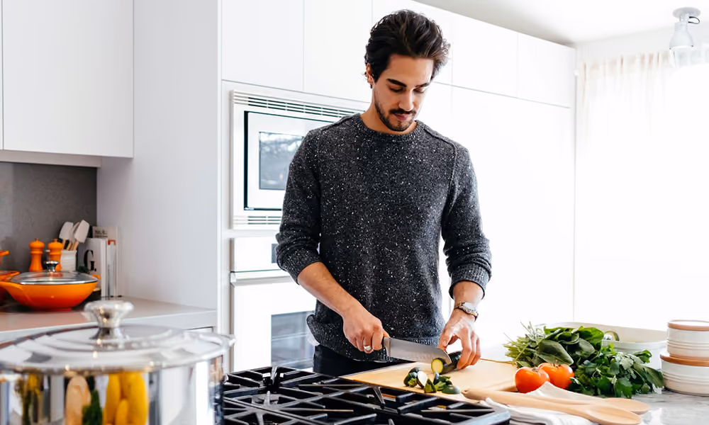 Man slicing zucchini on a cutting board in a bright home kitchen, with tomatoes and fresh herbs on the counter
