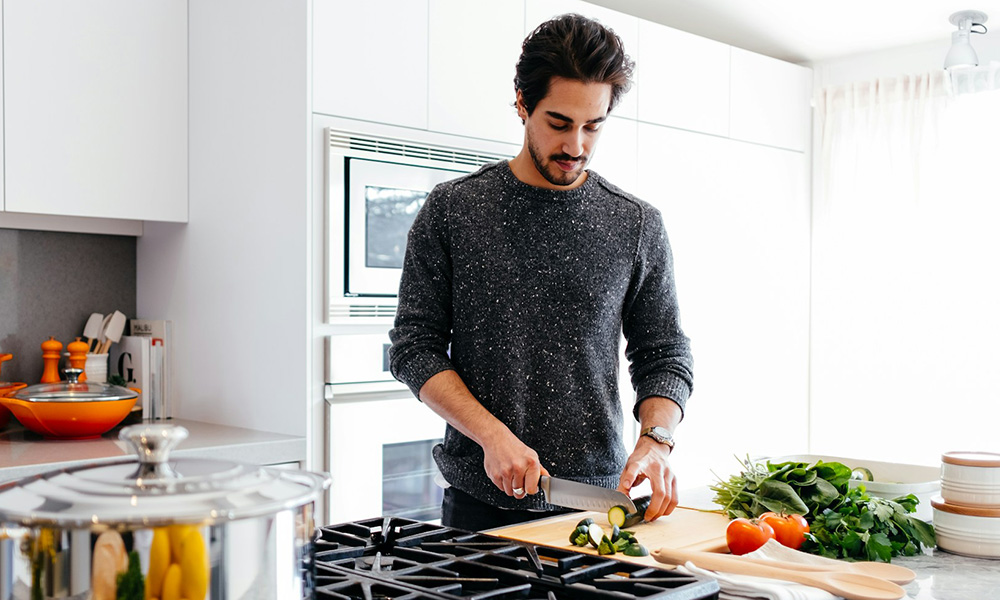 Man slicing zucchini on a cutting board in a bright home kitchen, with tomatoes and fresh herbs on the counter
