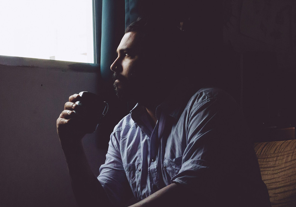 Bearded man sitting by a window holding a coffee mug, looking thoughtful