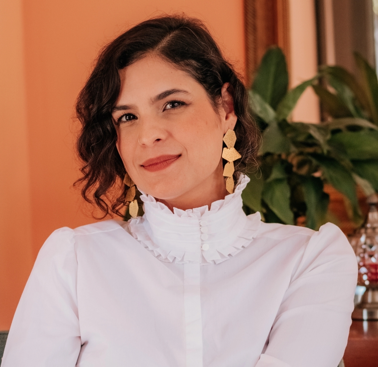 Woman with short curly hair wearing a white ruffled blouse and large gold earrings, against an orange background.