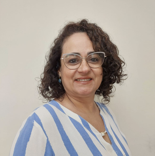 Woman with curly brown hair, glasses, and blue-striped top smiling against a beige background.