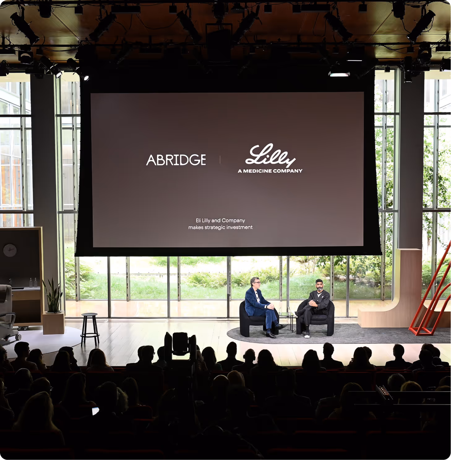 Two men seated and talking on a stage with Abridge and Lilly company logos on a large screen behind them.