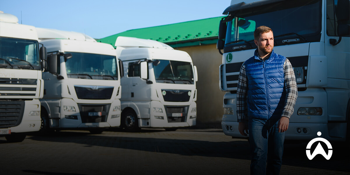 A person in a blue vest stands in front of several parked white trucks near a green-roofed building.