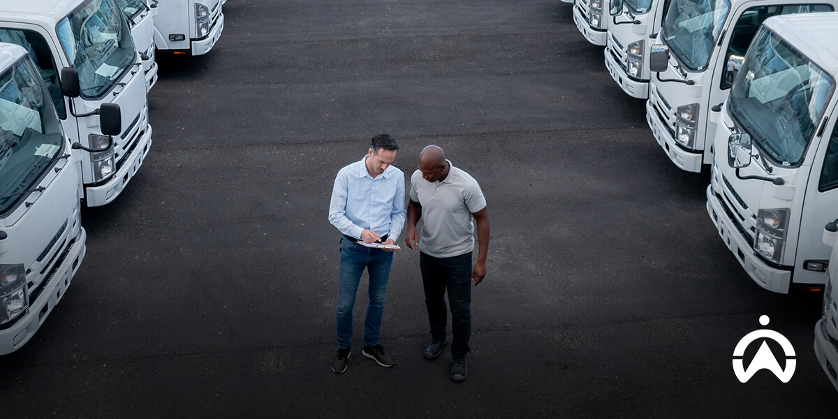 Two men stand on an asphalt lot surrounded by a fleet of white trucks, discussing details while holding a clipboard.