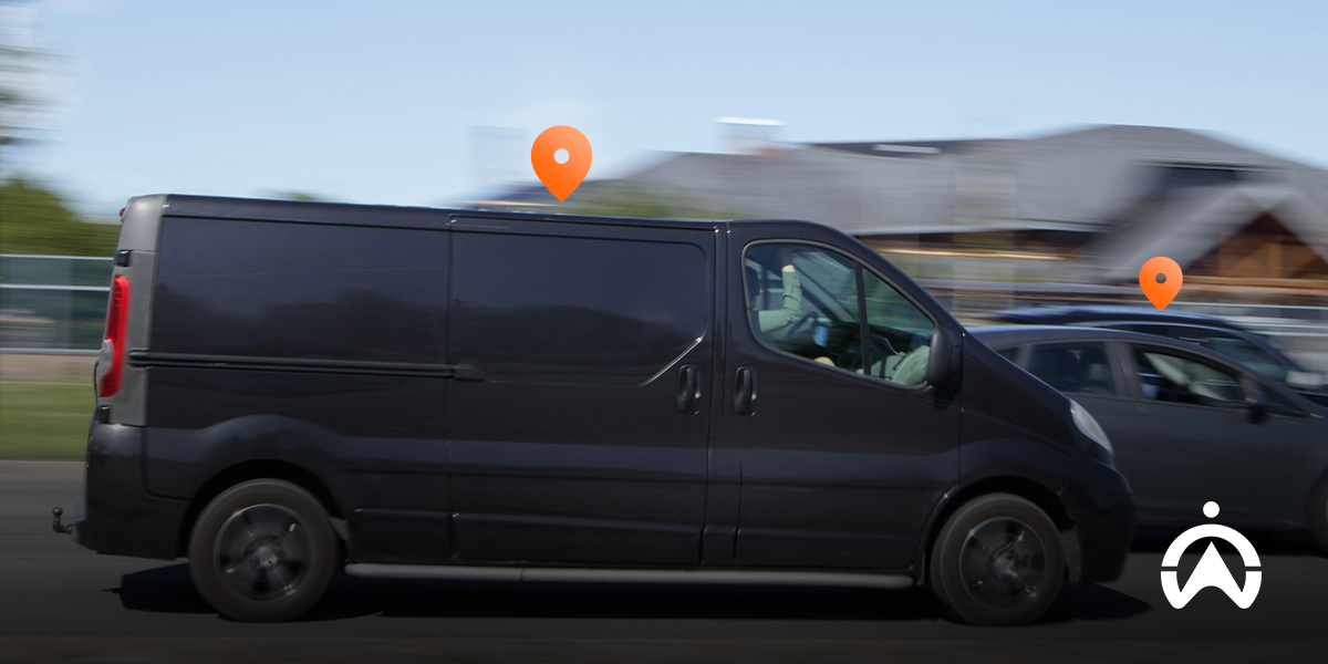 A black van speeds past, with orange location markers indicating its route against a blurred background of greenery and buildings.