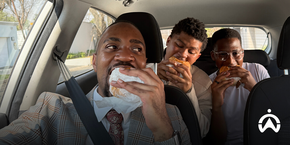 Three people in formal attire enjoying food in a car, with trees and a house visible outside the window.