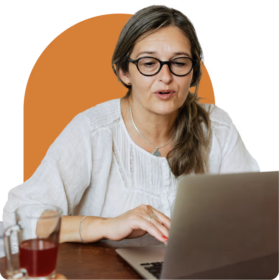 Middle-aged woman with glasses using a laptop at a desk, wearing a white blouse, with a glass of tea nearby.