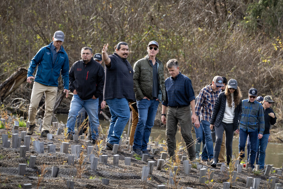 A young person carefully lights dry grass with a pitch stick under the guidance of an experienced fire practitioner, while other firefighters observe nearby in a forest clearing.