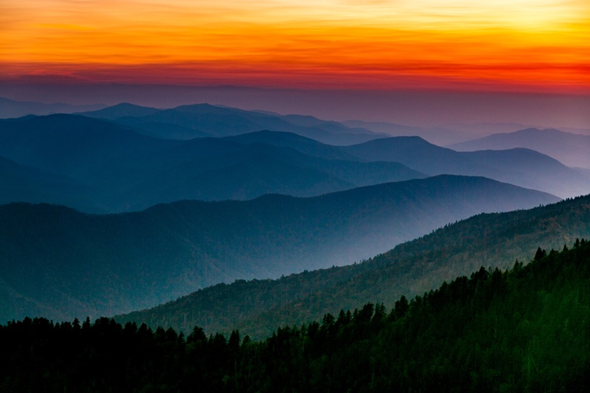 Sunrise over layered mountain ridges and forested slopes, showing the kinds of large, fire-adapted landscapes where forest and climate resilience work occurs.