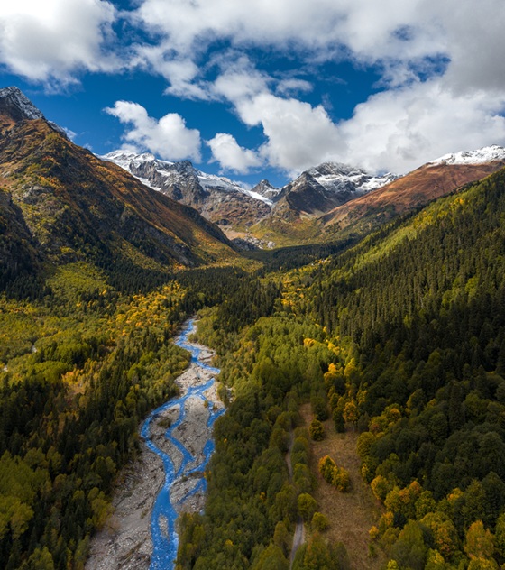 Aerial view of a river running through a forested mountain valley with snow-capped peaks and mixed fall colors, representing healthy, fire-adapted landscapes.