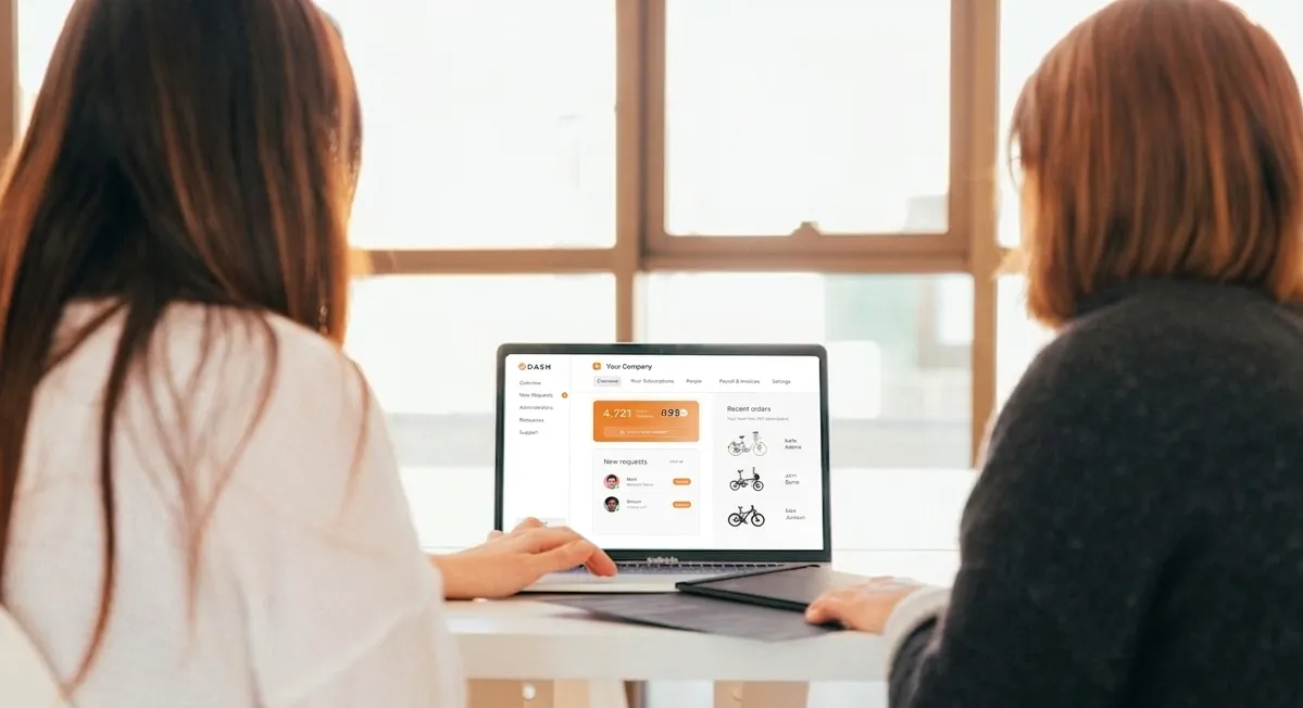 Two women seated at a table looking at a laptop screen displaying a company dashboard with orders and user requests.