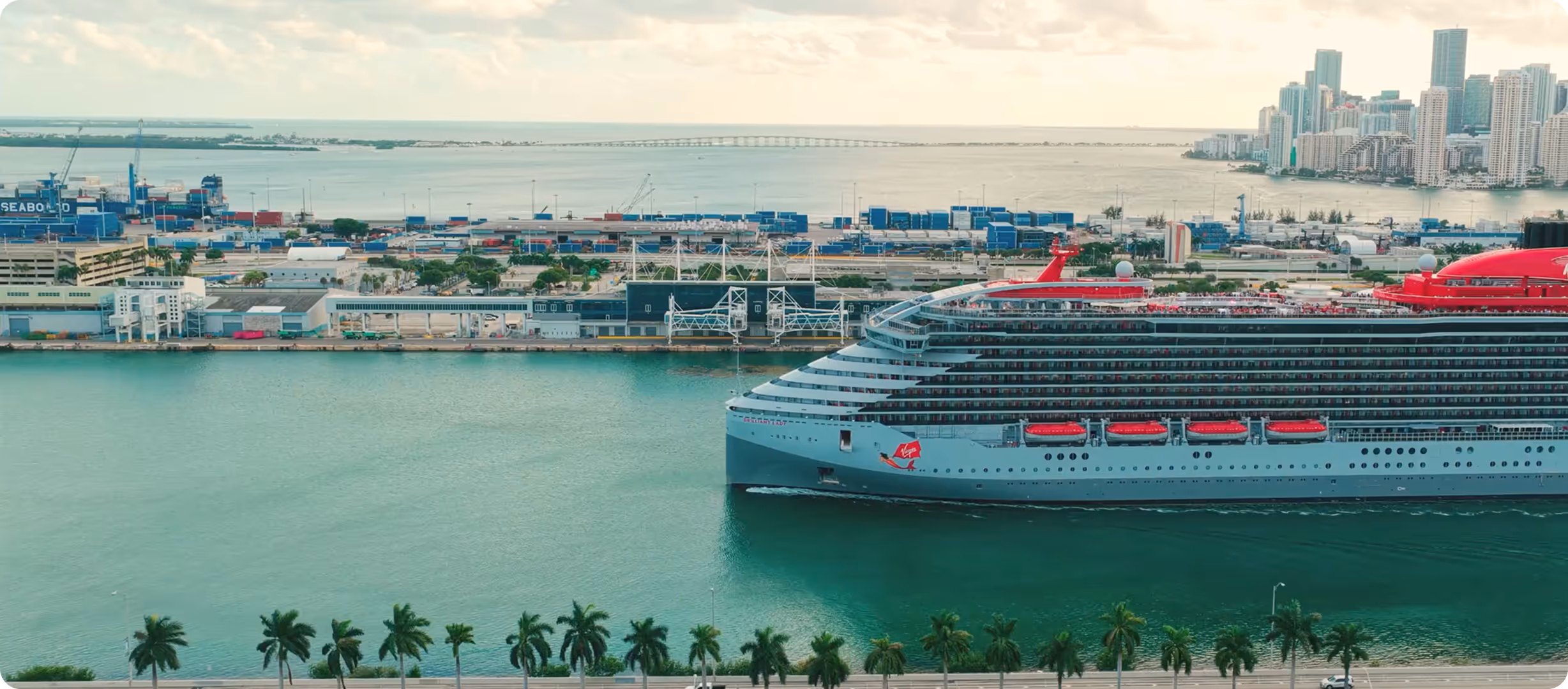 Large cruise ship with red lifeboats docked near a port with palm trees and city skyline in the background.