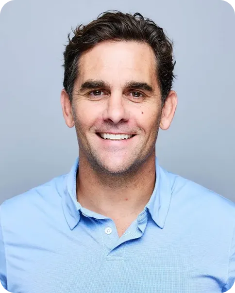 Smiling man with dark curly hair wearing a light blue collared shirt against a plain light gray background.
