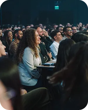 Audience of diverse adults smiling and attentively watching a presentation in a dimly lit conference or seminar room.
