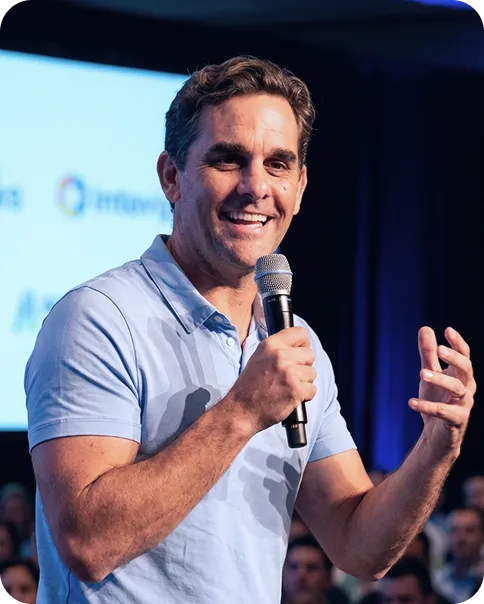 Smiling man holding a microphone and speaking at an indoor event with an audience in the background.