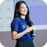 Smiling woman with long brown hair wearing a sleeveless black wrap dress against a black background.