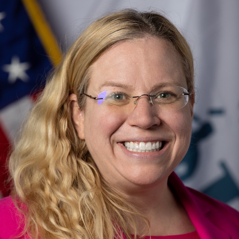 Smiling woman with long brown hair wearing a sleeveless black wrap dress against a black background.