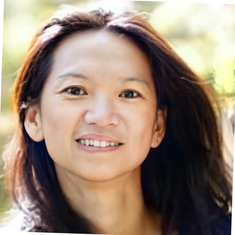 Smiling woman with long brown hair wearing a sleeveless black wrap dress against a black background.