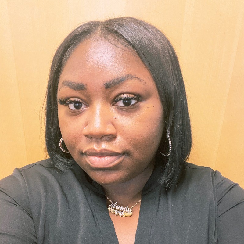 Smiling woman with long brown hair wearing a sleeveless black wrap dress against a black background.