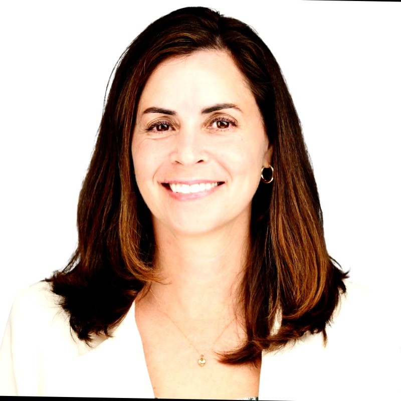 Smiling woman with long brown hair wearing a sleeveless black wrap dress against a black background.