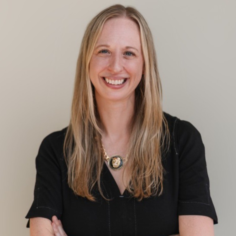 Smiling woman with long brown hair wearing a sleeveless black wrap dress against a black background.