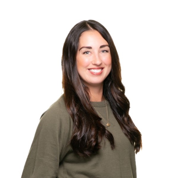 Smiling woman with long brown hair wearing a sleeveless black wrap dress against a black background.