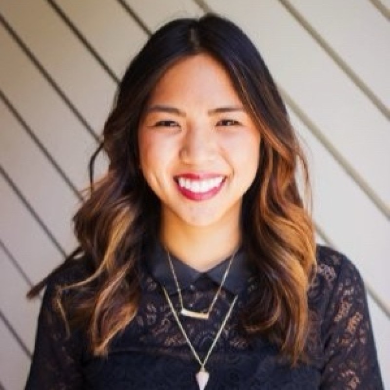 Smiling woman with long brown hair wearing a sleeveless black wrap dress against a black background.