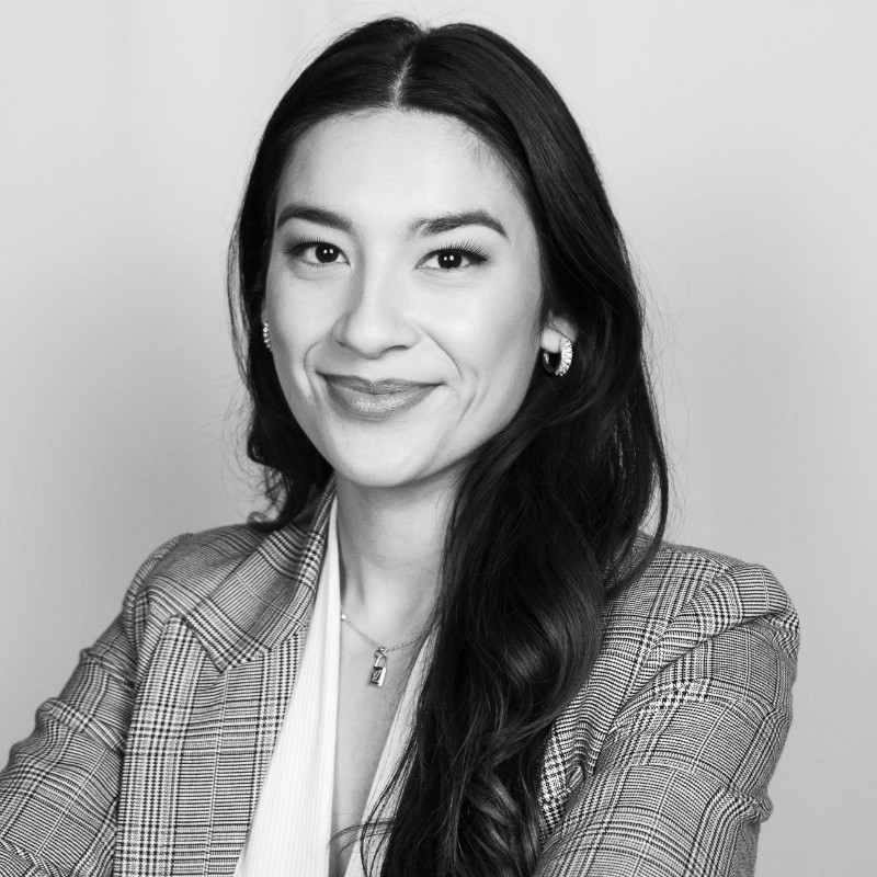 Smiling woman with long brown hair wearing a sleeveless black wrap dress against a black background.