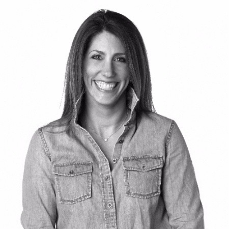 Smiling woman with long brown hair wearing a sleeveless black wrap dress against a black background.