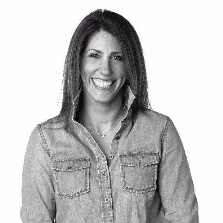 Smiling woman with long brown hair wearing a sleeveless black wrap dress against a black background.