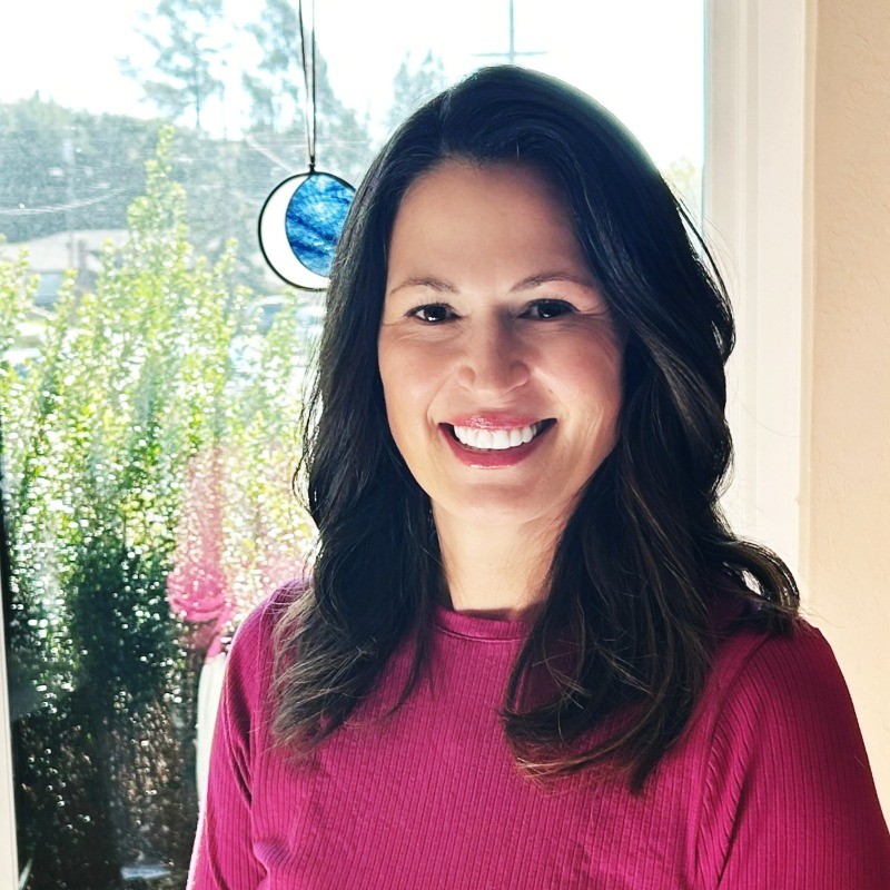 Smiling woman with long brown hair wearing a sleeveless black wrap dress against a black background.