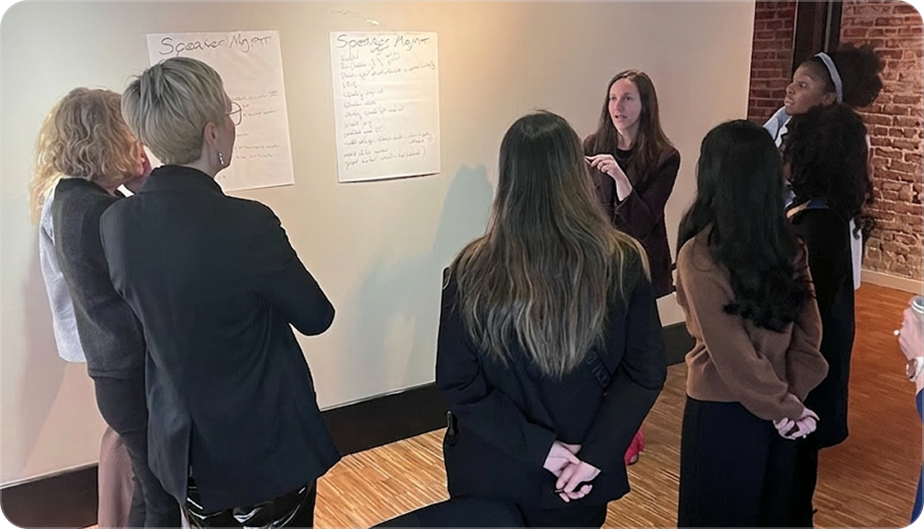 Group of seven people standing and listening to a woman explaining two large sheets of paper with handwritten notes on a wall in a meeting room.